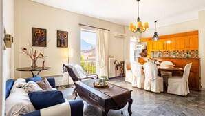 A living room with a sofa, a dining table with chairs, a kitchen area with wooden cabinets, and artwork on the walls. - Vicky s Cozy Nest Your Home by the Sea (Kalamata)