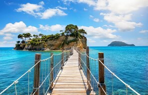 A wooden bridge leading to a rocky island with trees, surrounded by clear blue water.