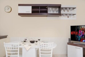 A dining area with a white table and chairs, a clock, a shelf with wine glasses, and a TV displaying the Netflix logo.