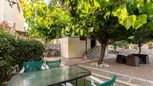 An outdoor patio area with a glass table and white chairs, surrounded by greenery and a stone wall. - Stoupa Oasis - Spitiko Garden Getaway (West Mani)