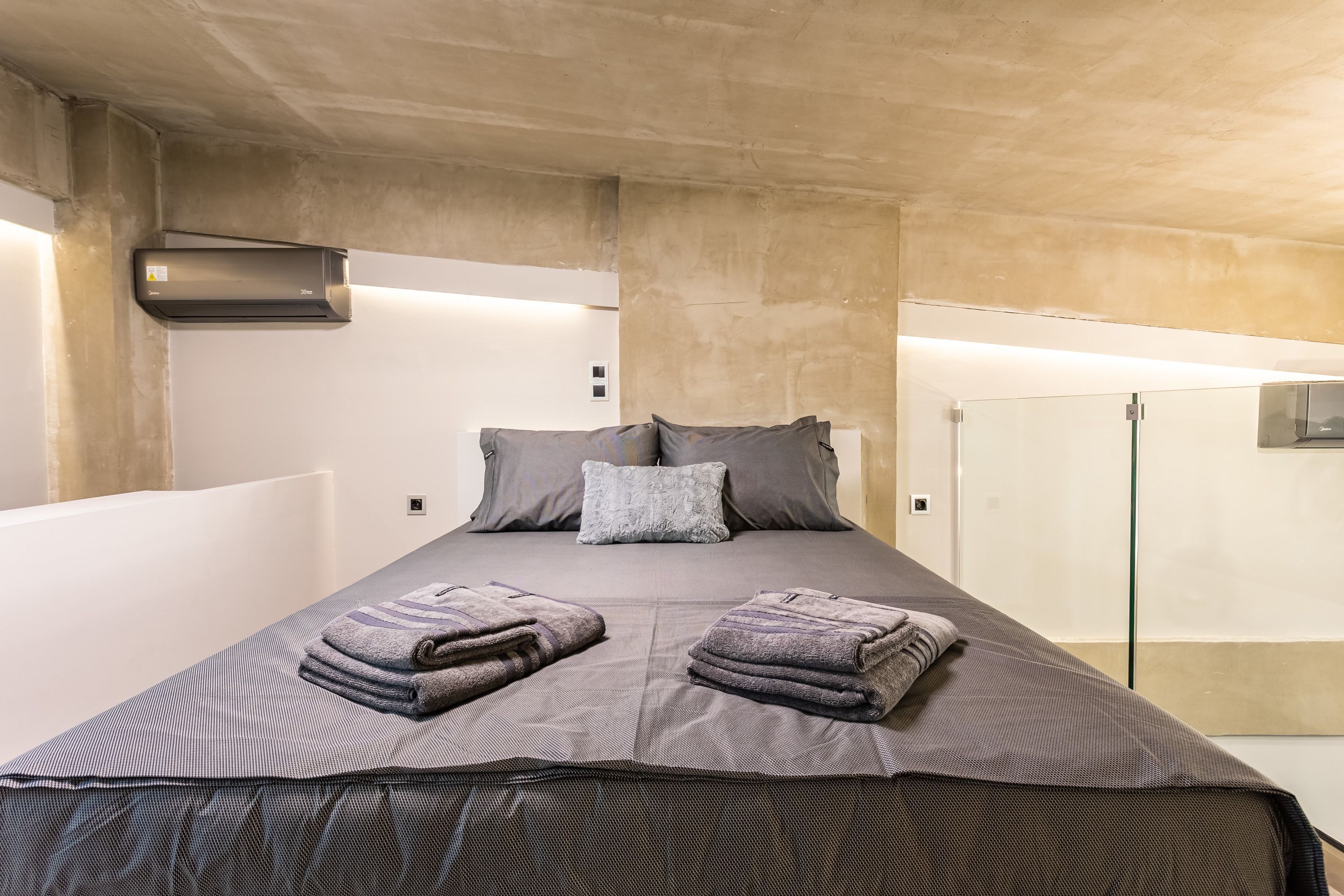 A modern hotel room with a large bed, grey bedding, and a built-in air conditioning unit.