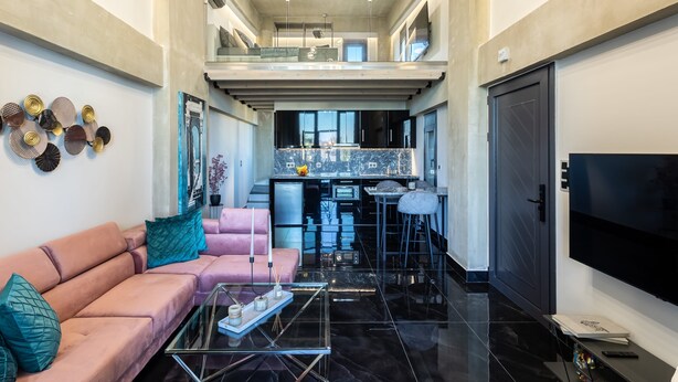 A modern living room with a pink sofa, a glass coffee table, and a kitchen area with a black countertop and a dining area. - Ventiri Lofts - Luxury Penthouse w Home Cinema (Kalamata)
