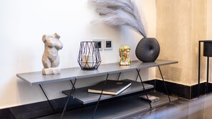 A modern interior with a grey console table displaying a stone sculpture, a candle in a wire holder, a decorative bust, and a tall vase with a feather. - Ventiri Lofts - Luxury Duplex w Private Garden (Kalamata)