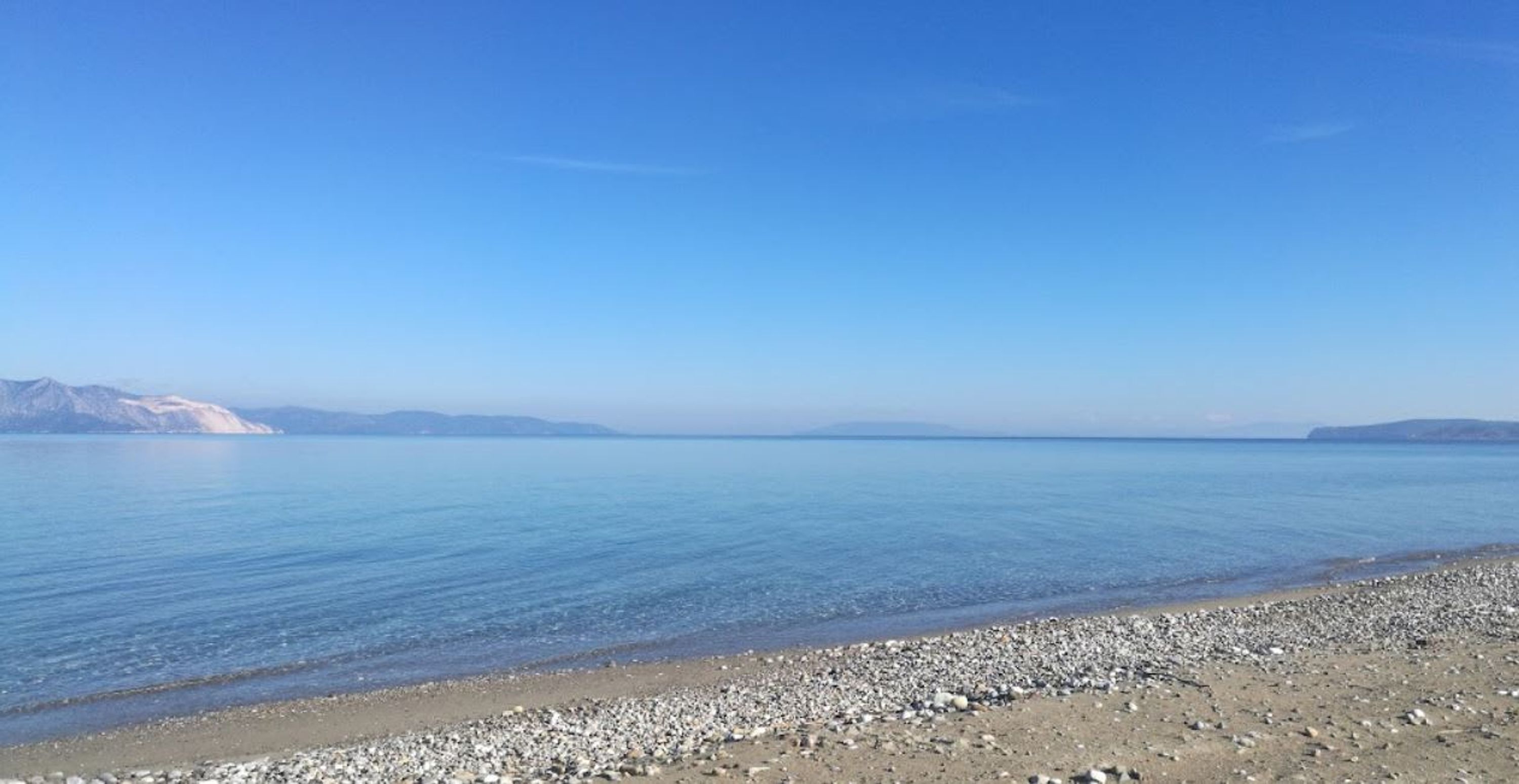 A clear view of a pebble beach with calm water and distant mountains under a blue sky.