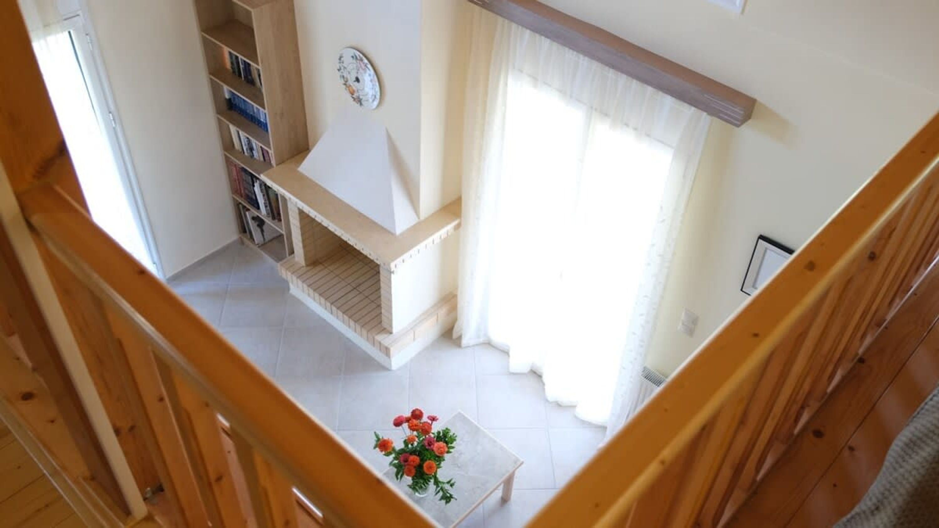 Une pièce avec un escalier en bois, une bibliothèque, une petite table ornée de fleurs et une fenêtre avec des rideaux.