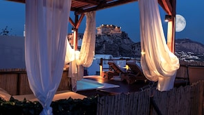 A rooftop terrace with a pool, lounge chairs, and a view of a historic castle under a full moon. - Santorini Luxury Suite w Rooftop Hottub Seaview (Santorini)
