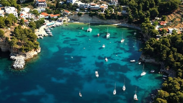 Une vue aérienne d’une zone côtière, avec des eaux turquoise limpides, de nombreux bateaux et un ensemble de bâtiments aux toits rouges.