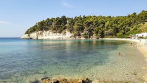 Une plage à l’eau claire, un rivage sablonneux et une colline boisée en arrière-plan.