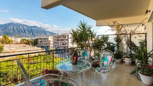 A balcony with a glass table and chairs, potted plants, and a view of a mountain and buildings. - Bright Comfy Flat Near the Beach the City Center (Kalamata)