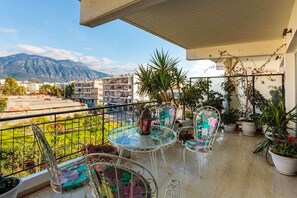 Un balcón con mesa y sillas de vidrio, plantas en macetas y vistas a la montaña y edificios.