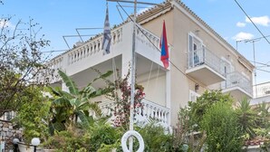 A building with a white facade, a balcony, and a flag. There are steps leading to the entrance, surrounded by potted plants and greenery. - Vibrant Spetses - Mata Colorful Nest (Spetses)