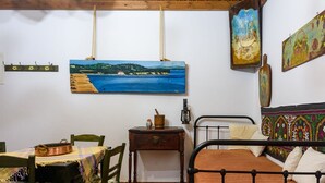 A room with a bed, a table with chairs, a small wooden nightstand, and a painting of a coastal landscape on the wall. - Eros Cozy Perfect Location Panoramic sea View (Pylos-Nestoras)