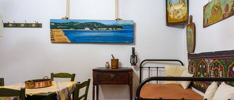 A room with a bed, a table with chairs, a small wooden nightstand, and a painting of a coastal landscape on the wall.