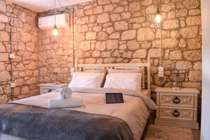 A bedroom with a stone wall, a bed with white bedding, two bedside tables, and a nightstand with a lamp. - Coastal Stone Hideaway With Stunning Scenery (West Mani)