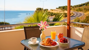 Un balcón con una mesa preparada para el desayuno, que incluye croissants, fruta y un vaso de zumo de naranja, con vistas a una carretera costera y al océano.