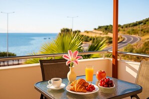 Un balcón con una mesa preparada para el desayuno, que incluye croissants, fruta y un vaso de zumo de naranja, con vistas a una carretera costera y al océano.