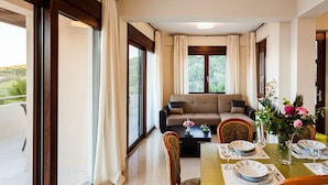 Un comedor con una mesa dispuesta para cuatro personas, un sofá al fondo y ventanas grandes con cortinas.