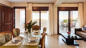 Un comedor con una mesa dispuesta para cuatro, sillas, una mesita de centro y vistas al mar a través de amplias ventanas.