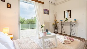 A hotel room with a bed, a bedside table with a lamp, a window with curtains, a radiator, a small table with a vase of flowers, and a view of the outdoors. - Seaside Family Haven - Avia s Spacious Getaway (West Mani)