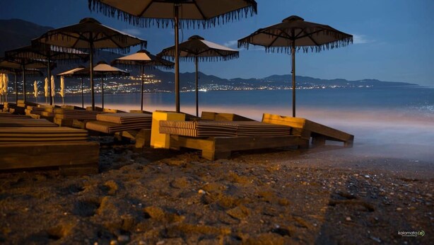 Beach umbrellas and wooden loungers on a pebbled beach at dusk with city lights in the background. - Stylish Seaside Home - Fully Equipped big Terrace (Kalamata)