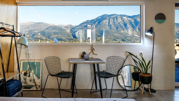 Quarto com uma grande janela com vista para as montanhas, uma mesa pequena com cadeiras, um abajur e uma planta em vaso.