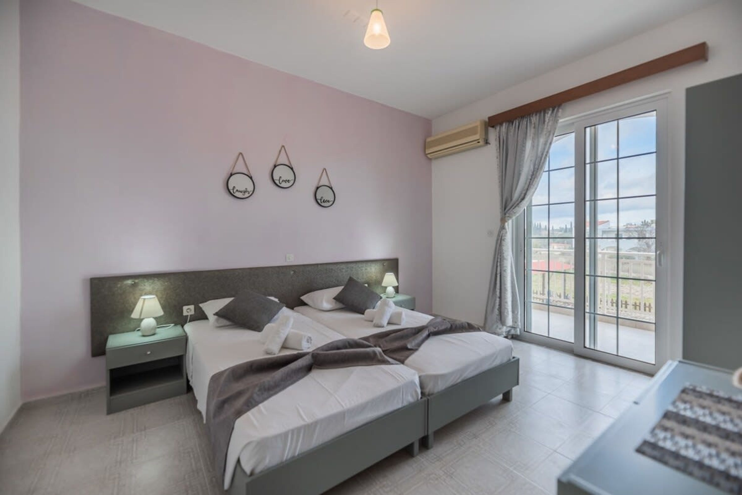 Een slaapkamer met twee bedden, een nachtkastje met een lamp, een wandplank met decoratieve objecten en een groot raam met gordijnen.