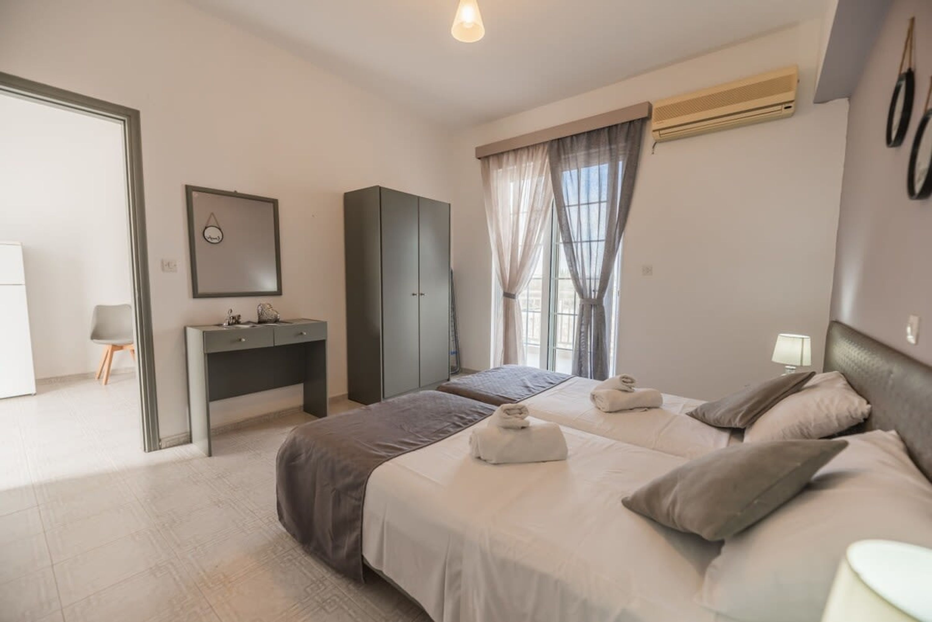 A modern hotel room with two beds, a desk, a wardrobe, and a large window with curtains.