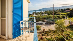Un balcon avec un volet bleu, une chaise blanche et une vue sur un paysage côtier, avec des maisons et des collines.