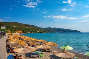 A beach with thatched umbrellas and beach chairs, a clear blue sea, and a hilly landscape in the background.