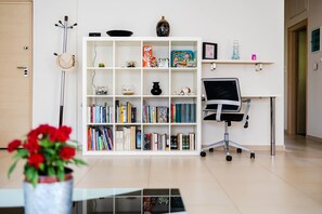 A modern office space with a white bookshelf filled with books and games, a desk with a chair, a vase with red flowers, and a coat rack. - Kalamata s Spacious Getaway-taygetos Amazing View (Kalamata)