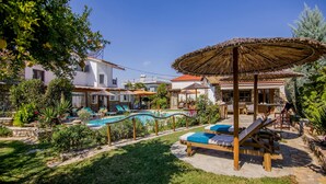 A pool area with lounge chairs and a thatched umbrella, surrounded by greenery and buildings.