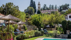 A villa with a swimming pool, lounge chairs, and a garden with a stone wall and trees.