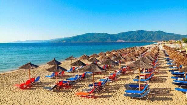 A beach with rows of sun loungers and thatched umbrellas, a clear blue sea, and mountains in the background.