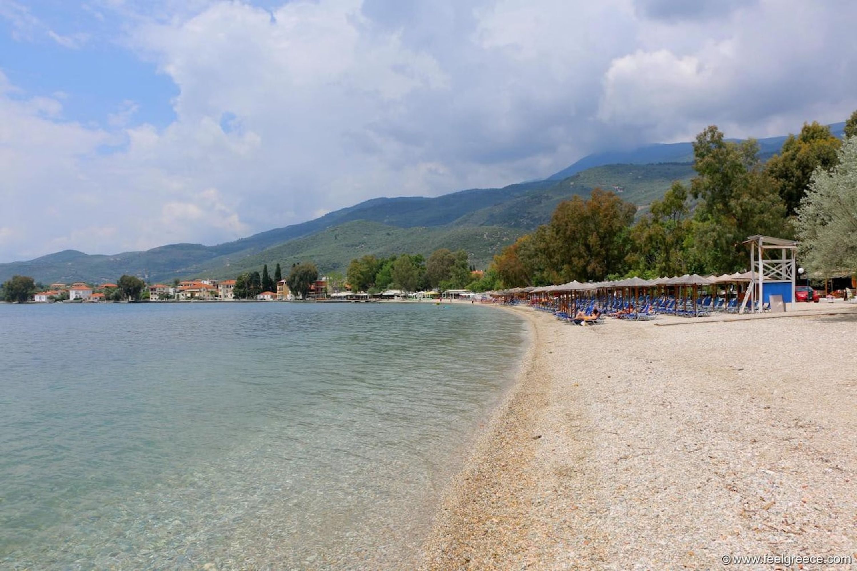 A pebbled beach with clear water, a row of beach umbrellas, and a mountainous backdrop.
