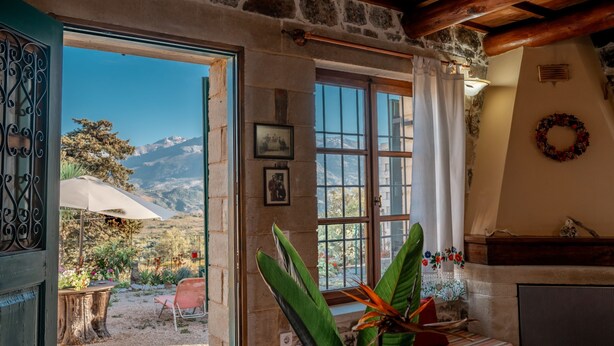 A stone-walled room with a wooden ceiling, a large window with curtains, a fireplace, and a view of mountains. - Maria s Serene Countryside Retreat by Chania (Chania)