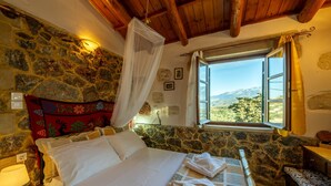 A bedroom with a stone wall, a bed with white linens, a bedside table with a lamp, and a window with a view of the countryside. - Maria s Serene Countryside Retreat by Chania (Chania)