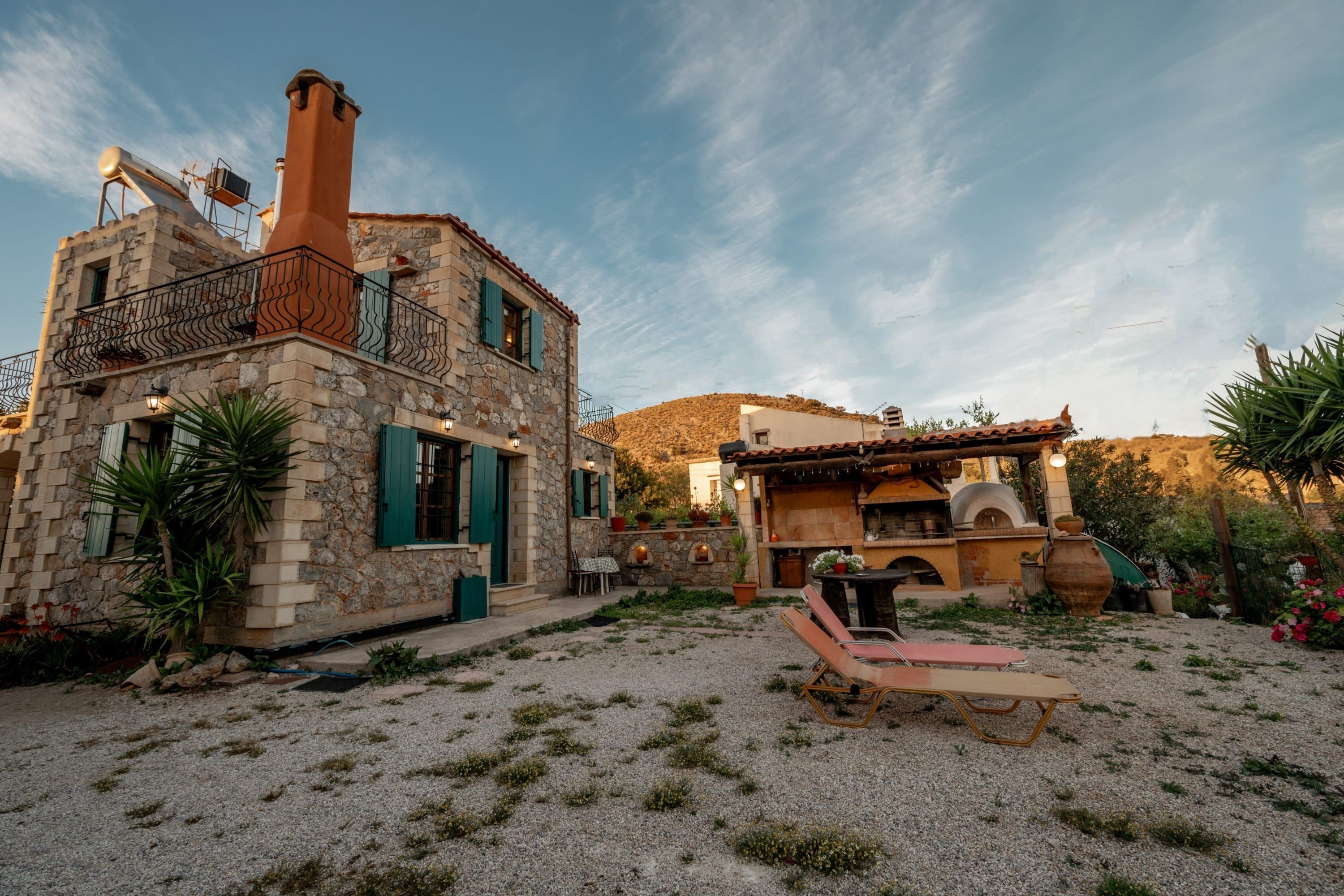 A stone building with a balcony, a grill area, and a lounge chair in a garden setting.