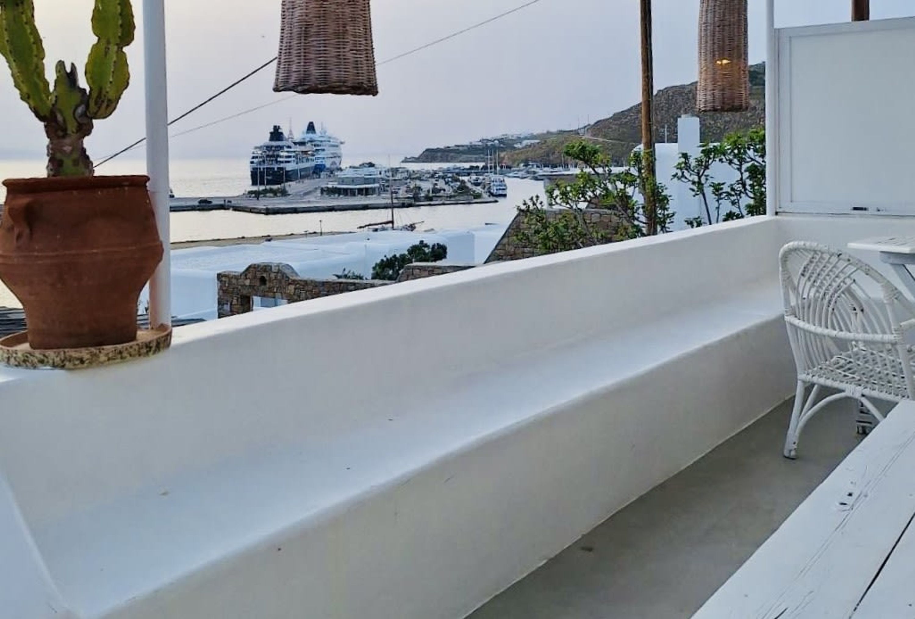 Een balkon met een witte rieten stoel, een potplant en uitzicht op een cruiseschip dat in een haven ligt, met boten en een bergachtig landschap op de achtergrond.