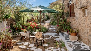 Un vialetto in pietra conduce a un patio con tavolo e sedie sotto un ombrellone verde, circondato da vasi con piante e fiori.