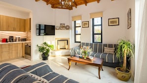 A cozy living room with a sofa, a coffee table, and a fireplace. There is a kitchen area with wooden cabinets and a television mounted on the wall. - Stoupa Stone Built Home A Breath Away to the Sea (West Mani)