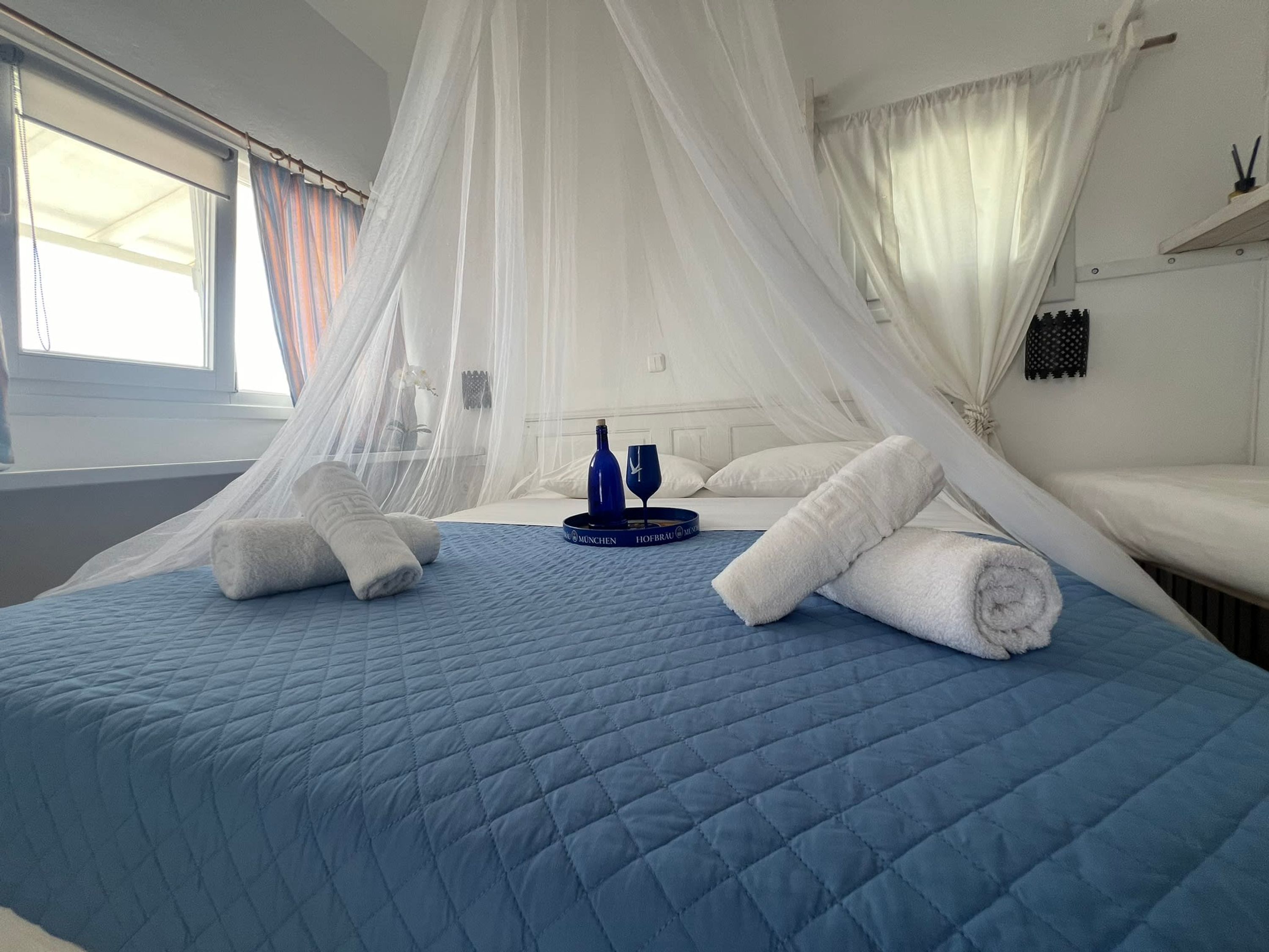 A hotel room with a bed covered in a blue quilt, white pillows, and a white canopy. There are two towels on the bed and a bottle of water on a tray.