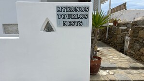 Ein Schild mit der Aufschrift 'MYKONOS TOURLOS NESTS', im Hintergrund eine Steinmauer und Kübelpflanzen.