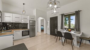 A modern kitchen with white cabinets, a built-in oven, and a stainless steel refrigerator. There is a dining area with a table and chairs, and a window with curtains. - Seaside Oasis Near Heraklion Villa Maro IV Suite (Hersonissos)