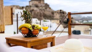 A wooden table with a fruit basket containing bananas, oranges, and a pineapple, and a glass of orange juice on a balcony overlooking a rocky landscape. - Santorini Rooftop Hot Tub Suite w Panoramic Views (Santorini)