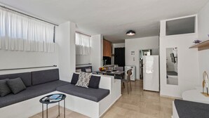 A modern apartment with a grey sofa, a small table, a kitchen area with a dining table, and a white refrigerator.