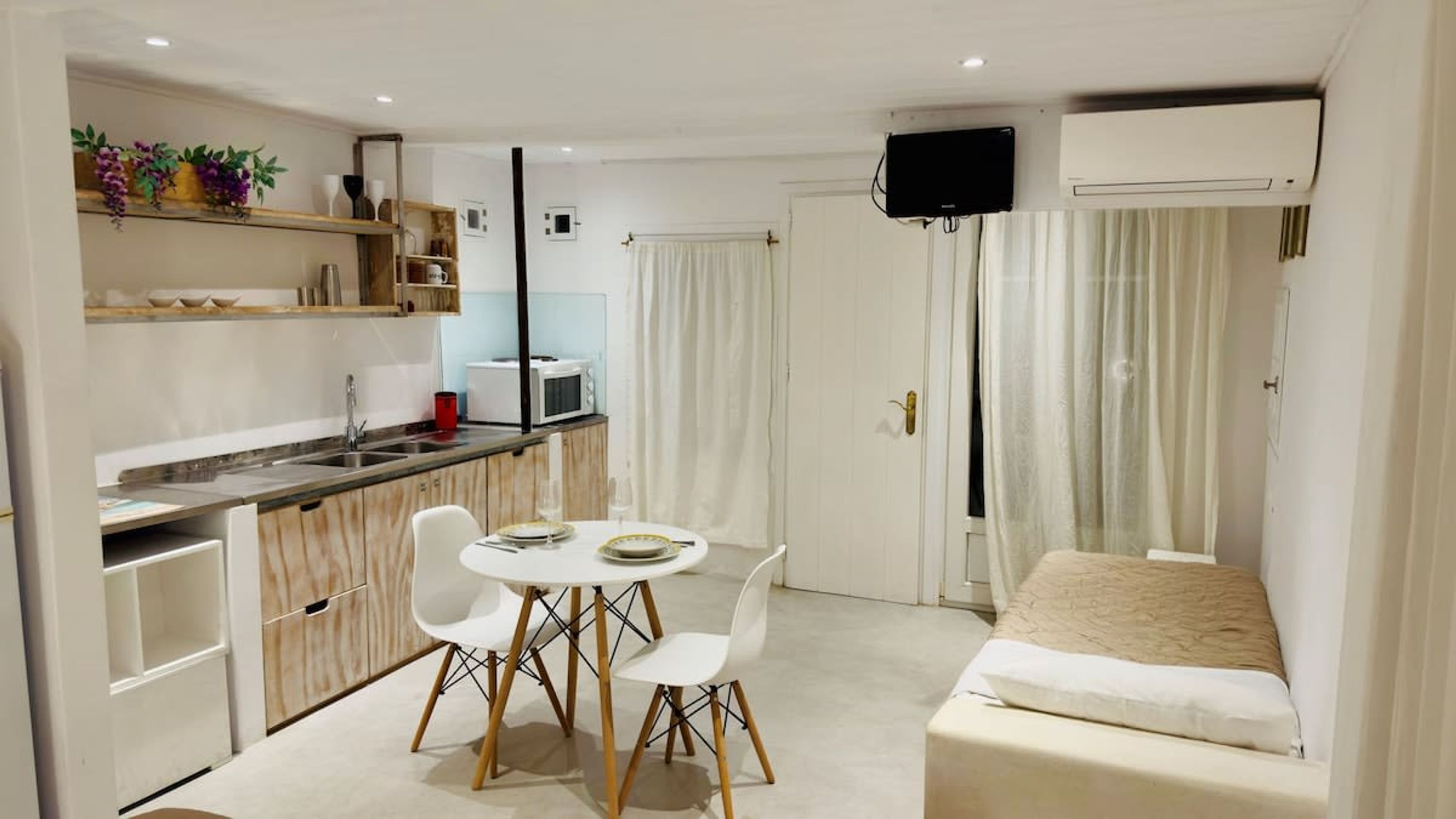 A compact hotel room with a kitchenette, a dining area with two chairs, a bed, and a TV mounted on the wall.