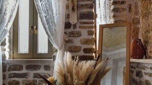 A room with stone walls, a bed, a wooden dresser, a mirror, and a window with curtains. - Lithos Messolongi Paradise - A Luxurious Retreat (Mesolongi)