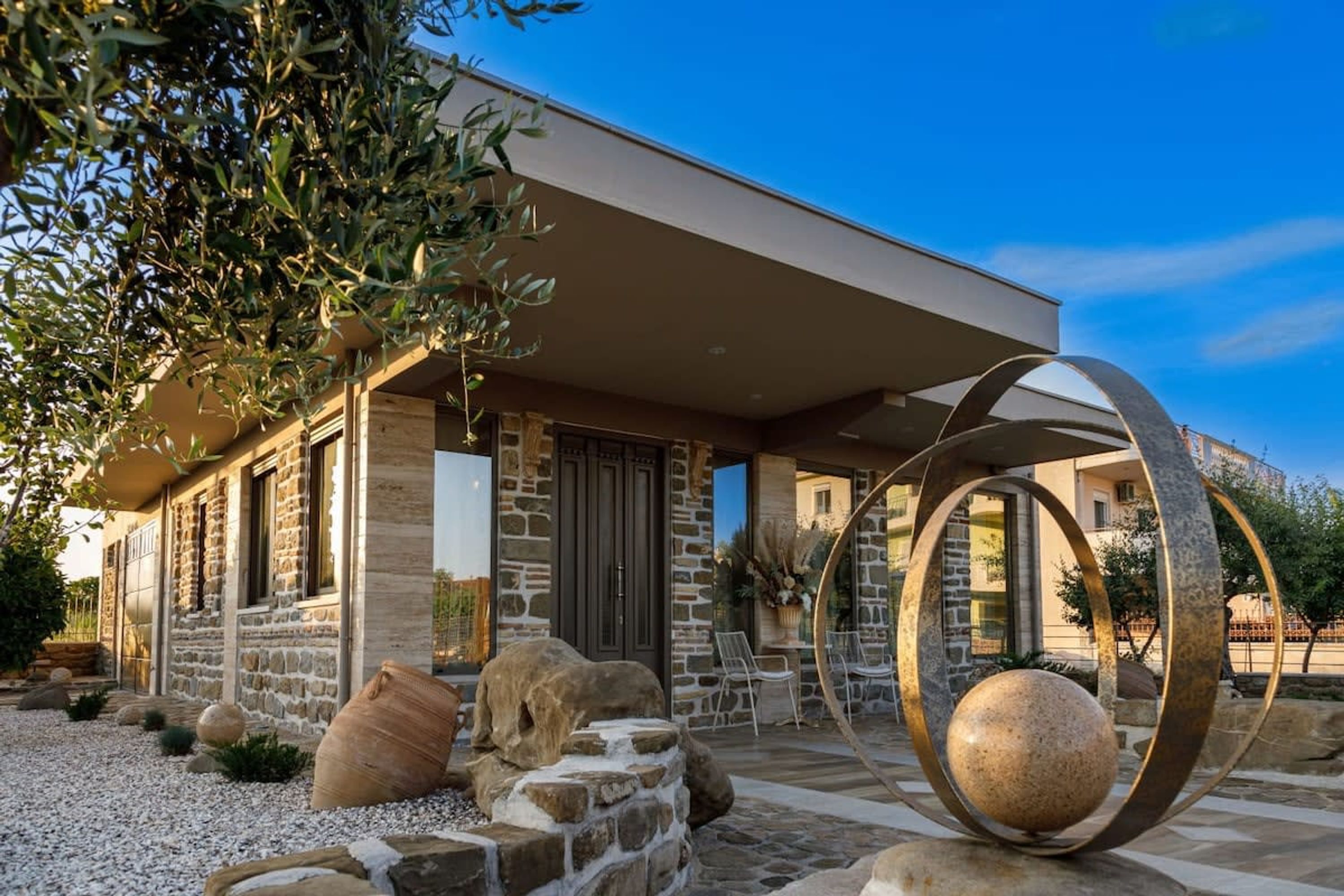 A modern house with stone walls, a large metal sculpture, and a gravel driveway.
