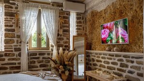 A room with a stone wall, a bed, a vanity with a mirror, and a TV on the wall. - Lithos Messolongi Paradise - A Luxurious Retreat (Mesolongi)