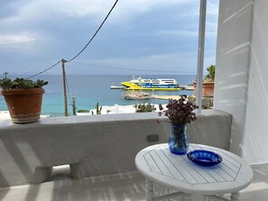 A balcony with a potted plant, a vase with flowers, and a table with a blue bowl. A ferry is visible in the background.